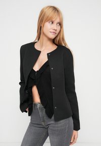 Μαύρο, στενό μπουφάν με στρογγυλή λαιμόκοψη και κούμπωμα με κουμπιά τσακίσματος. Το ύφασμα είναι λείο χωρίς σχέδια. Φοριέται πάνω από ένα μαύρο τοπ με βολάν και γκρι τζιν.