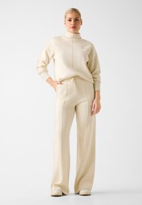 Loungewear-Set in Cremefarben mit einem Reißverschluss-Mockneck-Oberteil und weitem Hosenbein, versehen mit einem Tunnelzugbund und vertikalen Nähten.