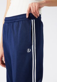 Marineblauwe trainingsbroek met witte zijsstrips, voorzien van een geborduurd logo op de zak en een elastische tailleband voor een verstelbare pasvorm.