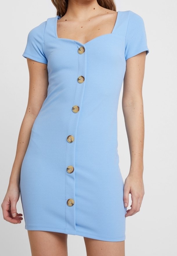 Femme portant une robe bleu clair ajustée avec des manches courtes et de gros boutons marron sur le devant, debout devant un fond uni.