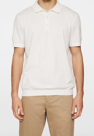 Mann trägt ein schlichtes weißes Polohemd mit kurzen Ärmeln und beige Hose, steht mit entspannten Armen seitlich am Körper vor weißem Hintergrund.