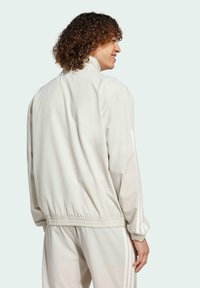 Giacca beige chiaro realizzata in materiale liscio. Presenta un colletto alto, polsini elastici e un orlo arricciato. Include sottili strisce bianche sulle maniche.