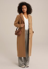 Brun lång kappa i ullblandning, med en spetsad krage och knäppning. Buren över en vit V-ringad topp och gråmönstrade byxor.