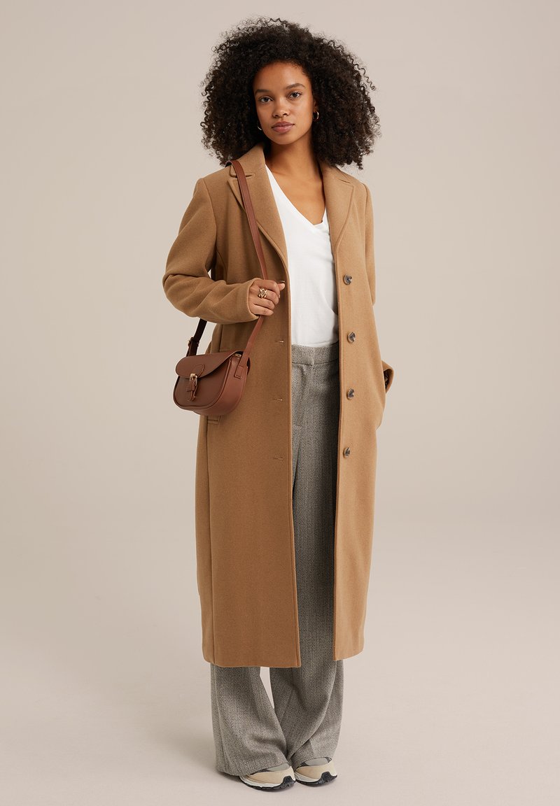 Brun lång kappa i ullblandning, med en spetsad krage och knäppning. Buren över en vit V-ringad topp och gråmönstrade byxor.