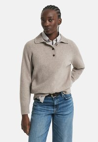 Una donna di colore con capelli intrecciati che indossa un maglione di lana beige sopra una camicia a righe e jeans blu, in piedi con una mano dietro la schiena.