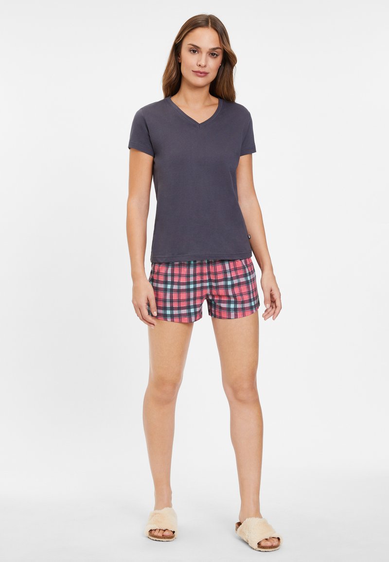 Camiseta de cuello en V de mangas cortas en gris oscuro combinada con pantalones cortos de cuadros en rosa y azul. El modelo lleva unas zapatillas de casa de peluche beige.