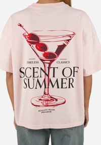 Lyserød oversized t-shirt med et rødt martiniglas-graphic og sort tekst, der læser "SCENT OF SUMMER" og "TIMELESS CLASSICS."