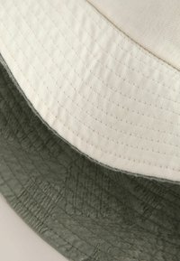 Kaksi kerrosta lippalakissa: ulkokerros on vaalea beige, teksturoitu kangas; sisäkerros on kuvioitu oliivivihreä, jossa on sileä pinta.