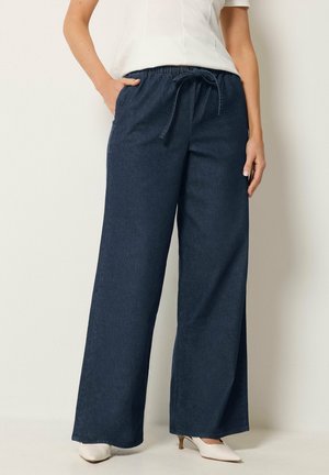 Donna che indossa pantaloni blu scuro a gamba larga con coulisse in vita, top bianco e scarpe bianche a tacco basso con punta, in piedi con una mano in tasca.