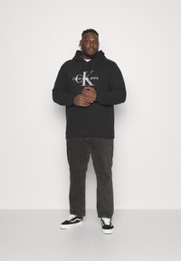 Black hoodie with "Calvin Klein Jeans" graphic, paired with charcoal jeans and black sneakers, set against a neutral background.