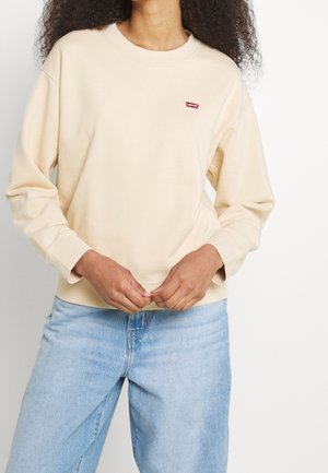 Personne portant un sweat Levi's couleur crème et un jean bleu clair, debout sur un fond blanc, mains ajustant le sweat.