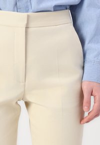 Bežīgas pielāgotas bikses ar gludu tekstūru, kurām ir šaurs piegriezums un plakana priekšpuse. Apvienotas ar gaiši zilu džinsu krekli.