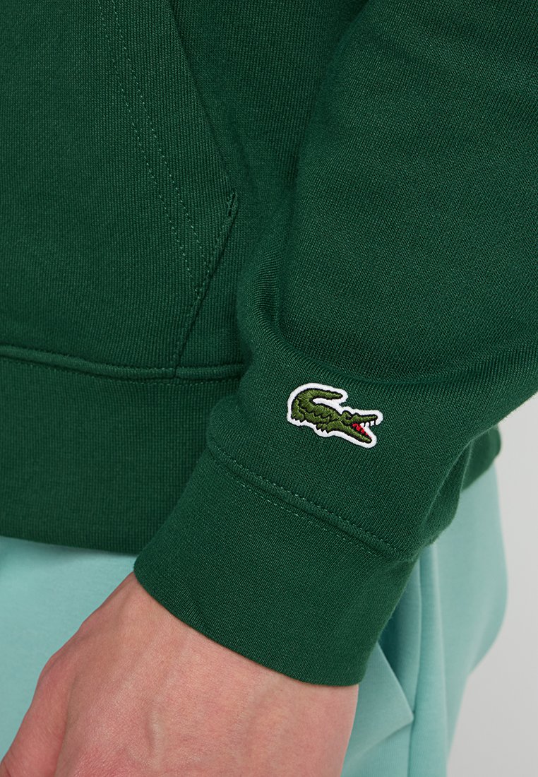Sweatshirt vert en tissu texturé, avec des poignets côtelés et un petit logo crocodile brodé sur la manche.