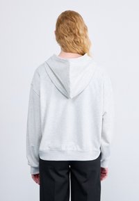 Sweat-shirt gris avec une capuche à cordon, coupe décontractée et poignets côtelés. Le tissu semble doux et légèrement texturé, avec une silhouette confortable.