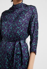 Abito di satin con base viola, caratterizzato da un motivo astratto di foglie verdi. Maniche lunghe e una cintura annodata in vita completano il design.