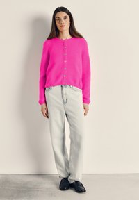 Moteris su ilgais tamsiais plaukais, vilkinti ryškiai rožinį mygtukais užsegamą kardiganą, šviesiai mėlynas džinsines kelnes ir juodus loafers, stovinti prie paprastos sienos.