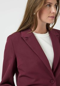 Blazer burdeos con solapa muesca, cierre de un solo botón y textura suave. Lleva puesto sobre una camiseta blanca, destacando su diseño estructurado.