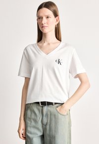 Biele bavlnené tričko s výstrihom do V, na ľavej strane hrude s logom, krátkymi rukávmi a voľným strihom. V kombinácii so svetlomodrými džínsami.