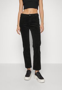 Pantalon en velours côtelé noir à coupe droite, avec une fermeture à boutons sur le devant, des passants pour ceinture et des poches latérales. Associé à un haut noir court et des baskets.