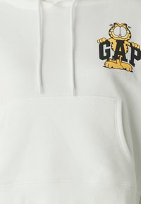 Sudadera blanca hecha de material suave, con un bolsillo canguro y un gráfico colorido de Garfield con "GAP" en letras negras en negrita.