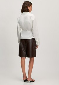 Witte blouse met lange mouwen en een cinched taille, gecombineerd met bruine leren shorts die tot op de knie komen. Zwarte instapsandalen maken de outfit compleet.