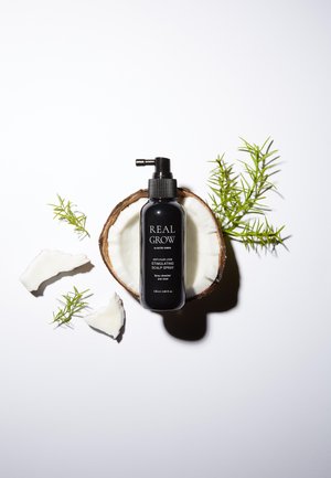 Bouteille de spray noire étiquetée "REAL GROW" sur une coque de noix de coco avec des morceaux de noix de coco blancs et des feuilles vertes, design minimaliste, forme ronde, 120 ml.