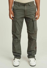 Cargo-broek in olijfgroen stof met meerdere cargozakken en zijzakken, voorzien van een knoopsluiting en een taps toelopende pasvorm. Vergezeld van witte sneakers.