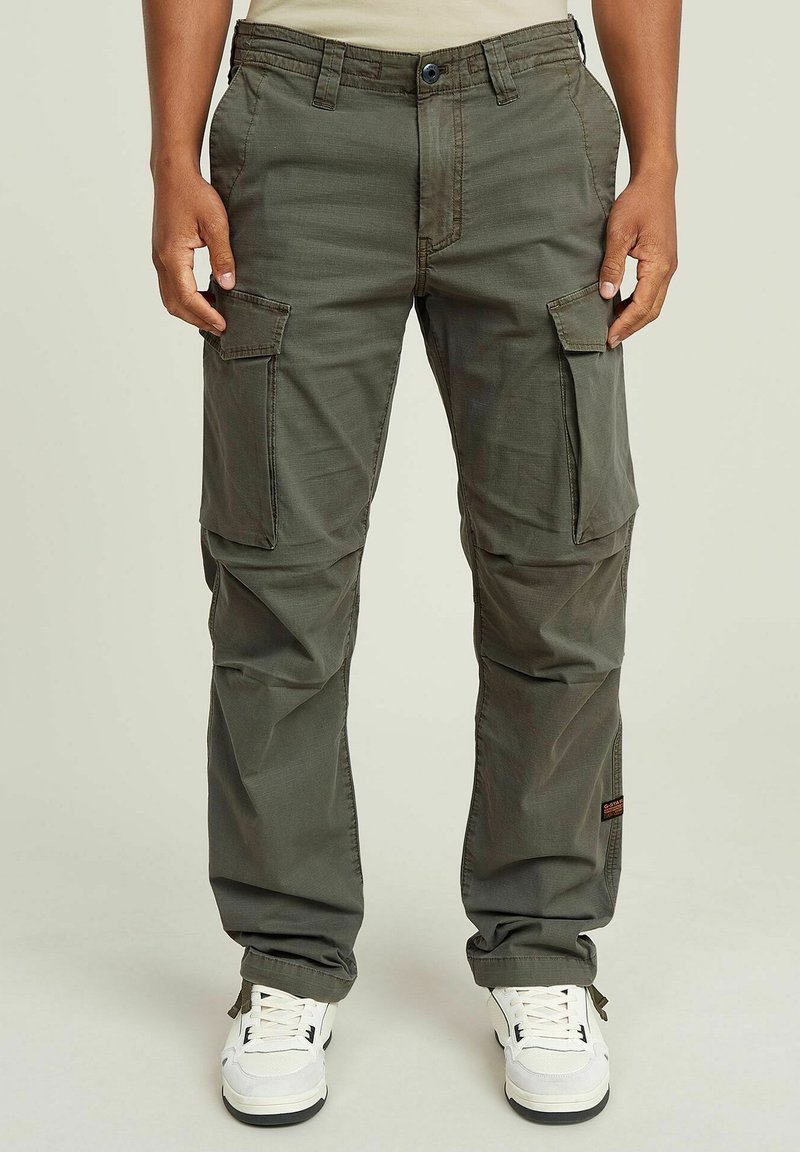 Cargo-broek in olijfgroen stof met meerdere cargozakken en zijzakken, voorzien van een knoopsluiting en een taps toelopende pasvorm. Vergezeld van witte sneakers.