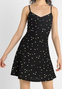 Czarna sukienka bez rękawów o dopasowanej górze i rozkloszowanej spódnicy, z kolorowymi kropkami w odcieniach różu, żółci i bieli na teksturowanym materiale.