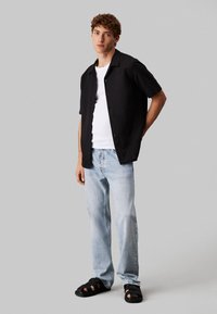 Camisa negra de manga corta tipo botón sobre una camiseta blanca, combinada con jeans rectos de color azul claro y sandalias negras de tiras. Fondo neutro.