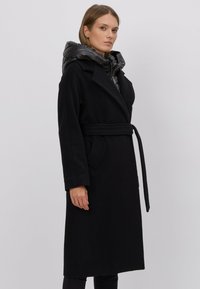 Czarny płaszcz z paskiem w talii, dużym kołnierzem i błyszczącym, pikowanym czarnym kapturem. Wykonany z miękkiego materiału, z długimi rękawami i bocznymi kieszeniami.