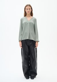 Maglia verde chiaro a maniche lunghe con scollo a V e dettagli plissettati, abbinata a pantaloni neri a gamba larga e vita alta realizzati in un materiale lucido.