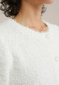 Vit texturerad jacka med en fluffig yta, med rund halsringning, sidofickor och två pärlknappar fram.