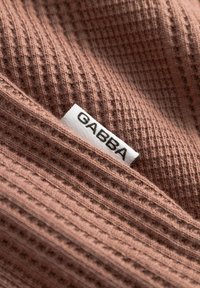 Hnědá látka s vaflovým pletením s bílou obdélníkovou visačkou na oblečení s nápisem "GABBA" černými velkými písmeny.