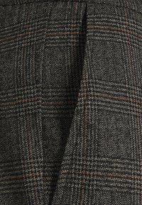 Tissu en tweed chevrons gris et noir avec un motif à carreaux rouge et blanc subtil, présentant un détail de couture pliée.