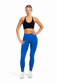 Czarny sportowy stanik z marszczeniami, zestawiony z gładkimi, wysokim stanem, intensywnie niebieskimi legginsami. Modelka ma na sobie białe buty sportowe.