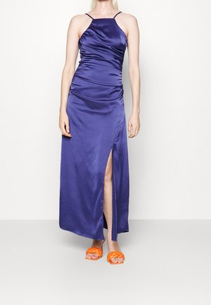 Kobieta w bezrękawnikowej, królewskim niebieskim satynowej sukience z rozcięciem do uda, w połączeniu z jasnopomarańczowymi, plecionymi sandałami na otwartym palcu.