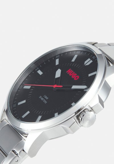 Montre en acier inoxydable argenté avec un cadran noir, logo "HUGO" rouge, grands index et couronne texturée, étanche jusqu'à 30 mètres.