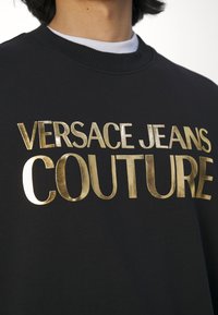 Černá mikina s kulatým výstřihem s velkým zlatým kovovým nápisem „Versace Jeans Couture“ přes hrudník.