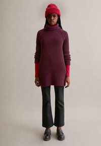 Gebreide coltrui in donkerpaars, gecombineerd met een langgebiesde rode top, zwarte flared pants en zwarte loafers; hoed in bijpassende paars-rode tint.