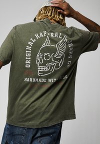T-shirt en coton vert olive avec un graphique de crâne usé présentant des ailes au dos, des lettres blanches et des accents rouges. Coupe décontractée.