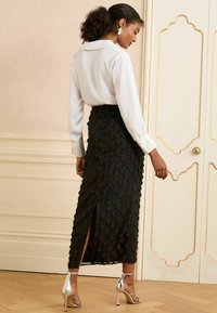 Jupe noire avec un motif floral texturé, fente sur le côté, associée à un chemisier blanc. Talons métalliques qui réfléchissent la lumière, ajoutant une touche brillante.