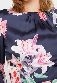Granatowa bluzka w kwiatowy wzór, z różowymi i białymi liliami. Gładki materiał z marszczonym detalem przy dekolcie, nadającym kształt.
