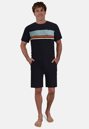 Junger Mann steht barfuß, trägt ein schwarzes T-Shirt mit blauen, orangefarbenen und weißen horizontalen Streifen sowie schwarze Shorts mit Vordertaschen.