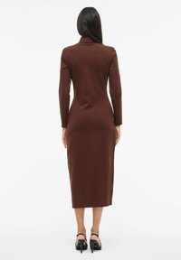 Ruskea, vartalonmyötäinen midimekko, jossa on pitkät hihat ja kaulus, valmistettu sileästä kankaasta. Yhdistetty mustien korkokengillä varustettujen sandaalien kanssa.