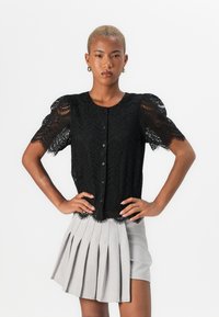 Blusa nera in pizzo con maniche a sbuffo corte e chiusura con bottoni, abbinata a una gonna plissettata grigia chiaro. Tessuto testurizzato con bordi scallop.