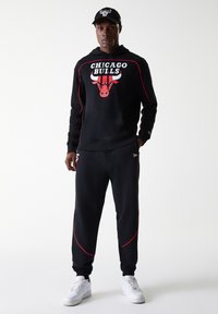 Conjunto de hoodie preto e calças de treino com o logótipo dos Chicago Bulls e linhas vermelhas de destaque, combinado com ténis brancos e um boné preto.