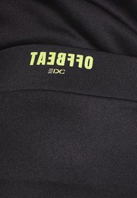 Tissu noir avec le texte jaune "OFFBEAT" et un texte blanc plus petit en dessous, visible en gros plan sur le matériau texturé.