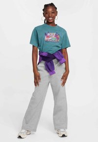 T-shirt de algodão azul-petróleo com um design gráfico colorido, combinado com calças de fato de treino cinzentas e um suéter roxo amarrado à cintura. Ténis prateados.