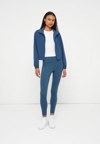 Casaco leve azul com colarinho alto e fecho éclair, combinado com leggings justas azuis. Ténis brancos completam o visual.